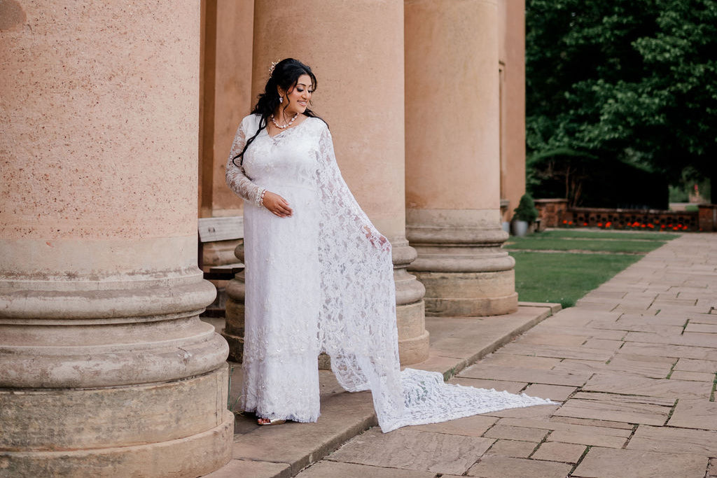 A woman wearing a long white lace saree stands between large stone columns in an outdoor garden setting, holding out the sheer lace train of the saree.