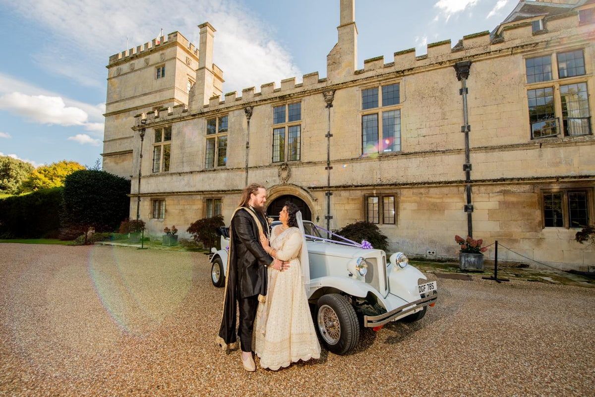 Sherni London bride and groom in fusion church wedding ceremony wearing white chikankari lucknowi lehenga and black asymmetric sherwani with gold buttons and trim standing in front of vintage car