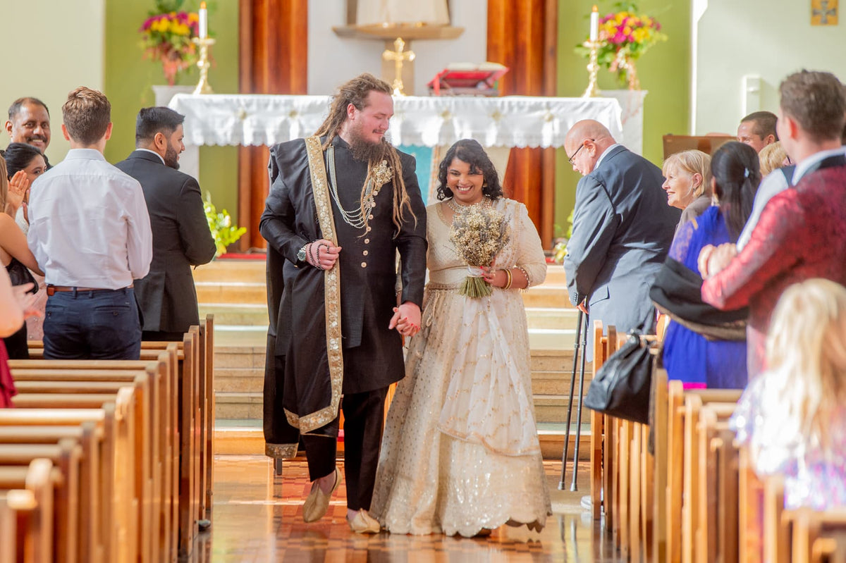 Sherni London bride and groom in fusion church wedding ceremony wearing white chikankari lucknowi lehenga and black asymmetric sherwani with gold buttons and trim