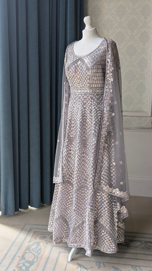 A heavily embellished silver-gray anarkali gown with intricate gold and silver embroidery, displayed on a white mannequin in an indoor setting with blue curtains and patterned wallpaper.