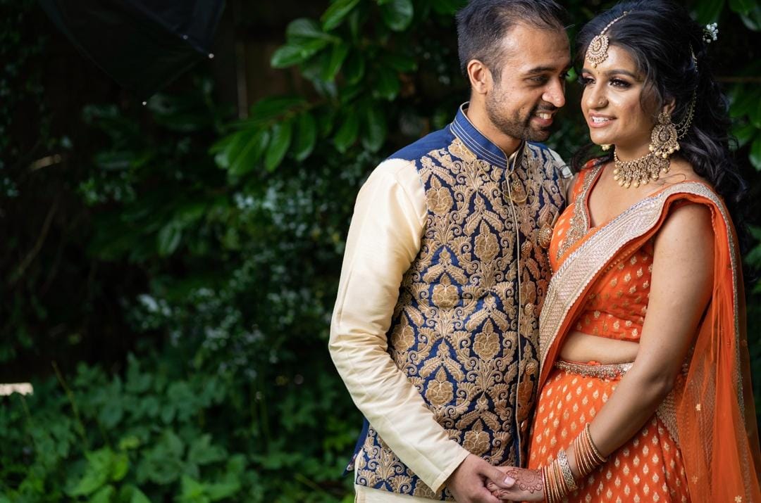 Couple in traditional Indian attire with bride in orange lehenga from Sherni London standing close together with a green leafy background